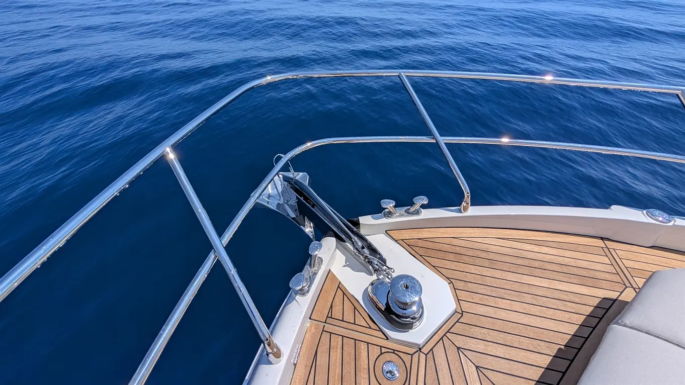 View of a boat's bow with polished railings, wooden deck, and anchor mechanism against a calm blue sea.